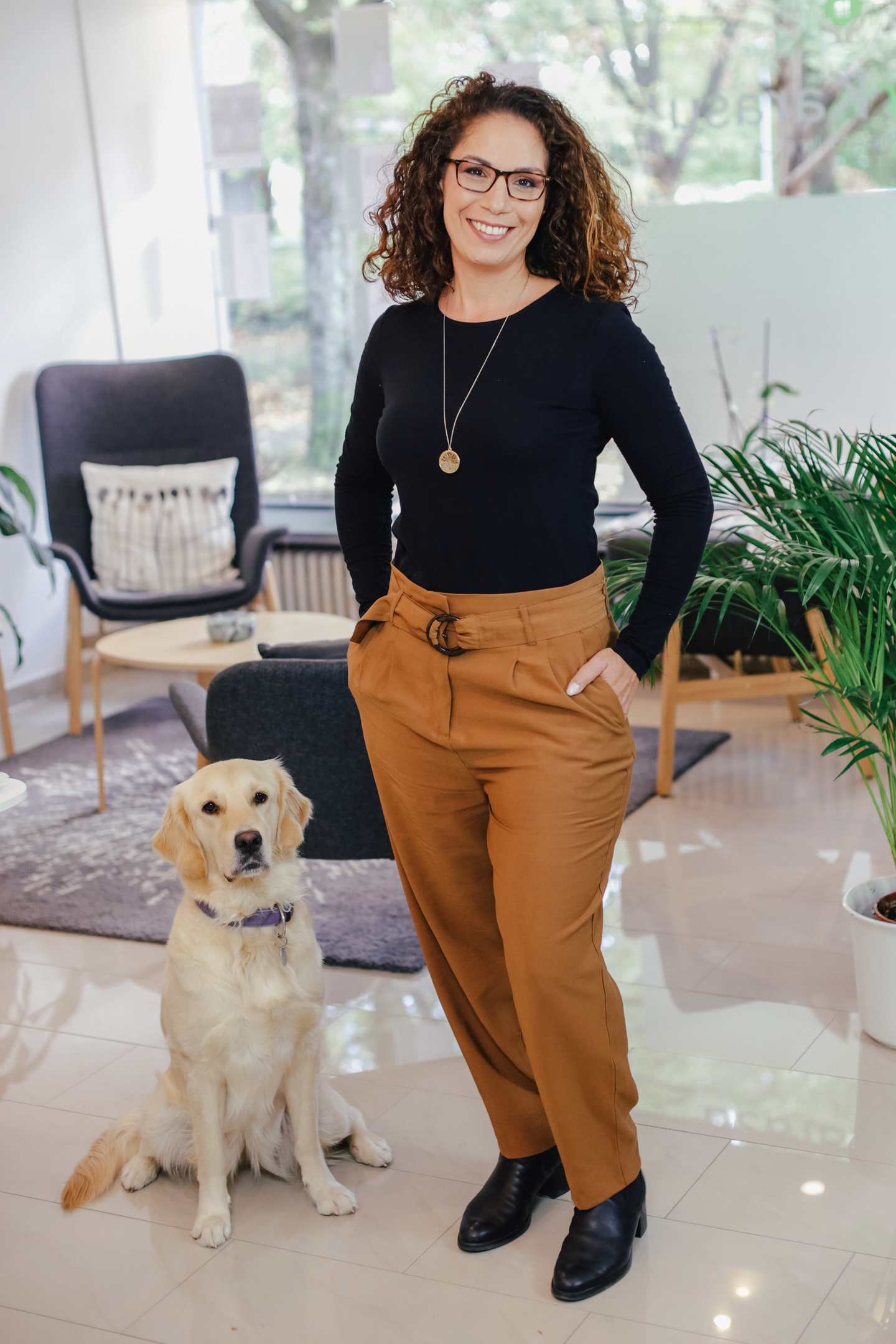 Eine Frau in schwarzer Bluse und braunen Hosen steht lächelnd neben einem goldenen Labrador, der auf dem Boden sitzt, in einem modernen, hellen Raum mit Pflanzen und Möbeln im Hintergrund.