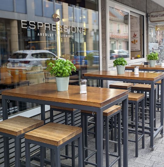 Außenbereich eines Cafés mit hölzernen Tischen und Stühlen, Pflanzen und Schildern im Hintergrund.