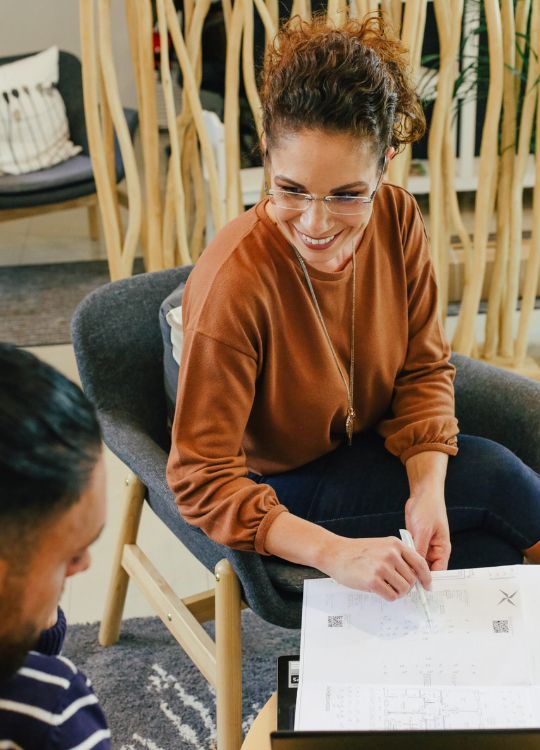 Eine Frau in einem braunen Oberteil sitzt lächelnd und schreibt auf einem Blatt Papier, während sie mit jemandem spricht.