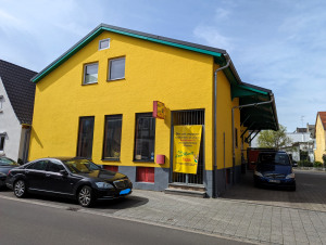 Ein gelbes Haus mit grünem Dach steht an einer Straße, vor dem Eingang sind ein Auto und ein Lieferwagen geparkt.