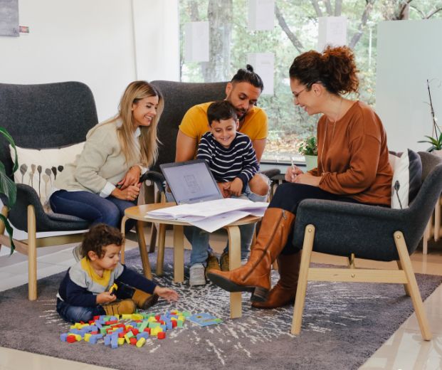 Eine Familie sitzt in einem Wohnzimmer, während ein Kind spielt und ein Erwachsener an einem Laptop arbeitet.